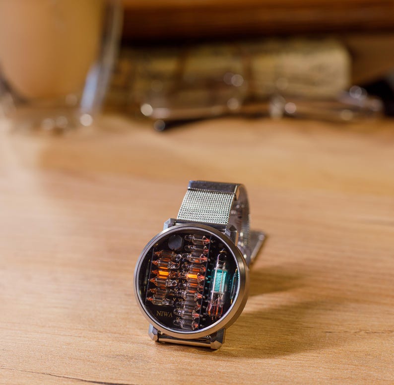K&ouml;nnte beinhalten: Eine runde Armbanduhr mit einem silberfarbenen Metall-Mesh-Armband und einem dunklen, kreisf&ouml;rmigen Zifferblatt. Das Zifferblatt zeigt freiliegende elektronische Komponenten, darunter mehrere kleine Glasr&ouml;hren mit leuchtend orangefarbenen und gr&uuml;nen Elementen. Das Wort NIWA ist in Wei&szlig; auf der unteren linken Seite des Zifferblatts aufgedruckt.