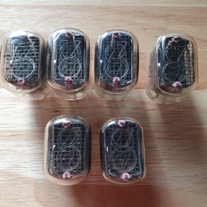 Peut inclure: Sept tubes Nixie vintage avec des boîtiers en verre transparent et un treillis métallique interne. Chaque tube a une broche rose en haut. Les tubes sont disposés sur une surface en bois brun clair.