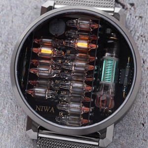 K&ouml;nnte beinhalten: Eine runde Armbanduhr mit silberfarbenem Metall-Mesh-Armband und einem dunklen Zifferblatt mit freiliegenden elektronischen Bauteilen. Das Zifferblatt zeigt den Schriftzug NIWA und enth&auml;lt mehrere kleine Glas-Vakuumr&ouml;hren, einige leuchten orange.