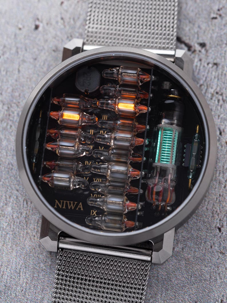 K&ouml;nnte beinhalten: Eine runde Armbanduhr mit silberfarbenem Metall-Mesh-Armband und einem dunklen Zifferblatt mit freiliegenden elektronischen Bauteilen. Das Zifferblatt zeigt den Schriftzug NIWA und enth&auml;lt mehrere kleine Glas-Vakuumr&ouml;hren, einige leuchten orange.