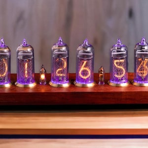 May include: A digital clock with six glass tubes displaying numbers in a warm orange glow. The tubes are mounted on a dark wood base with purple lighting and gold accents. A unique, retro-style timepiece.