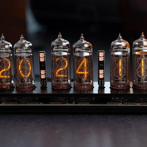 Könnte beinhalten: Eine Digitaluhr, die "2024" in leuchtend orangefarbenen Ziffern anzeigt. Die Uhr verfügt über sechs Glasröhren mit aufwendigen Innendesigns, die auf einer schwarzen Basis montiert sind. Das Design der Uhr erinnert an Vintage-Technologie.