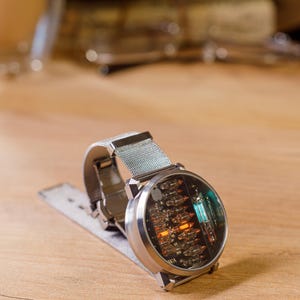 K&ouml;nnte beinhalten: Eine silberfarbene Armbanduhr mit einem runden Geh&auml;use und einer sichtbaren internen Nixie-R&ouml;hrenanzeige. Die Uhr verf&uuml;gt &uuml;ber ein Metall-Mesh-Armband und ein klares Glaszifferblatt, das leuchtende orange und blaue Elektronikbauteile zeigt.