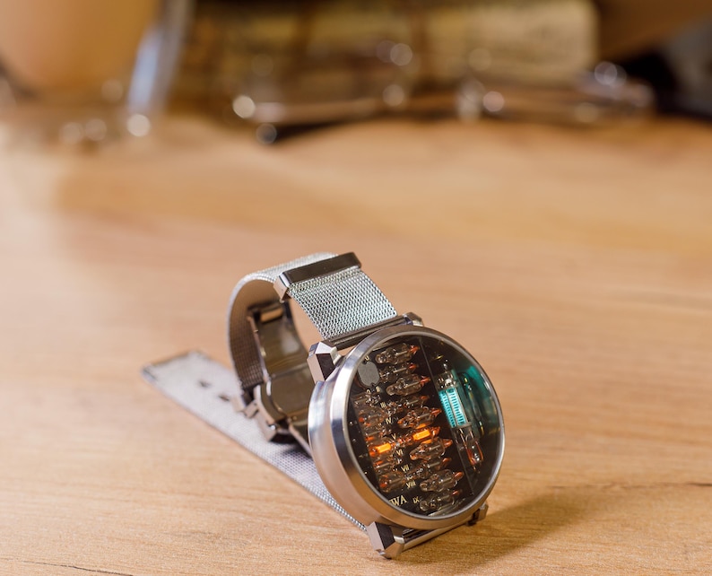 K&ouml;nnte beinhalten: Eine silberfarbene Armbanduhr mit einem runden Geh&auml;use und einer sichtbaren internen Nixie-R&ouml;hrenanzeige. Die Uhr verf&uuml;gt &uuml;ber ein Metall-Mesh-Armband und ein klares Glaszifferblatt, das leuchtende orange und blaue Elektronikbauteile zeigt.