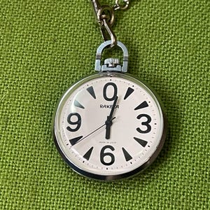 Peut inclure: Une montre de poche Raketa argentée avec un cadran blanc, des chiffres arabes noirs aux positions 0, 3, 6 et 9, et des aiguilles noires. La montre présente une couronne de remontage en haut, une chaîne en métal et l'inscription MADE IN USSR.
