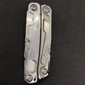 Op de afbeelding: Een zilveren multitool met de merknaam "LEATHERMAN Rev" zichtbaar. De tool is gesloten en toont zijn zijkant met diverse uitsparingen en schroefkoppen. De tool ligt op een donkere ondergrond.