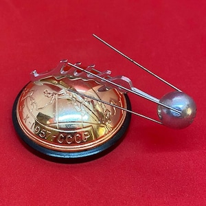 以下が含まれることがあります： ヴィンテージの人工衛星スプートニクのピンバッジ。金色の地球儀に地図と「X-1957-CCCP」の文字が描かれています。ピンバッジには、2本のアンテナが付いた銀色の衛星と、地球儀の上に装飾的な銀色の要素があります。赤い背景に配置されています。