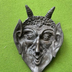 Könnte beinhalten: Eine dunkelgraue Schale aus Gussmetall in Form eines stilisierten Teufelsgesichts. Das Objekt zeigt zwei spitze Hörner, große Ohren, eine markante Nase und einen breiten, grinsenden Mund. Die Oberfläche hat ein verwittertes, texturiertes Finish.