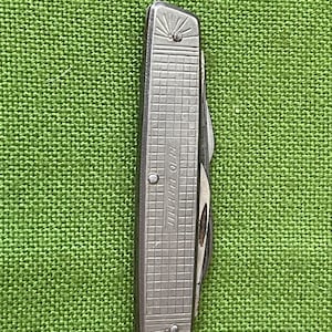 May include: A closed silver-toned pocket knife featuring a textured grid pattern on the handle. The metal surface displays a sunburst engraving at both ends and the text "J.A. HENCKELS" embossed in the centre of the rectangular grid design.