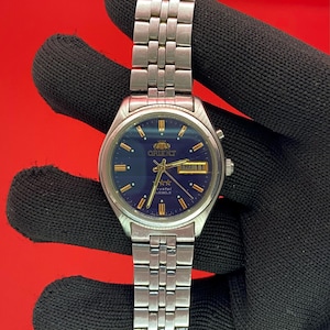 Könnte beinhalten: Eine silberfarbene Orient-Uhr mit blauem Zifferblatt und silbernem Armband. Das Zifferblatt zeigt den Tag und das Datum an. Die Uhr wird vor einem roten Hintergrund gehalten.