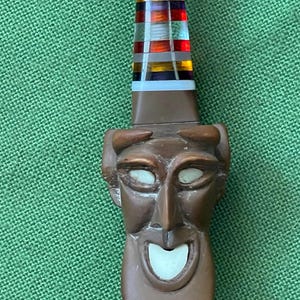 Peut inclure: Un manche sculpté marron représentant un visage stylisé avec des yeux et une bouche incrustés de blanc. Au-dessus du visage, le manche se rétrécit en une section cylindrique ornée de bandes horizontales en verre ou résine rouge, jaune et bleu.