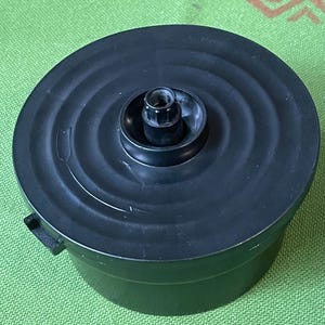May include: A black circular plastic food processor lid featuring a central raised hub with a gear-like attachment point. The lid has a smooth, slightly concave surface with concentric circular ridges and a small tab on the side for locking.