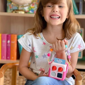 May include: A child holds a pink and white toy radio with red knobs and a small screen. The radio has a colourful beaded strap. The child wears a white t-shirt with colourful star graphics.