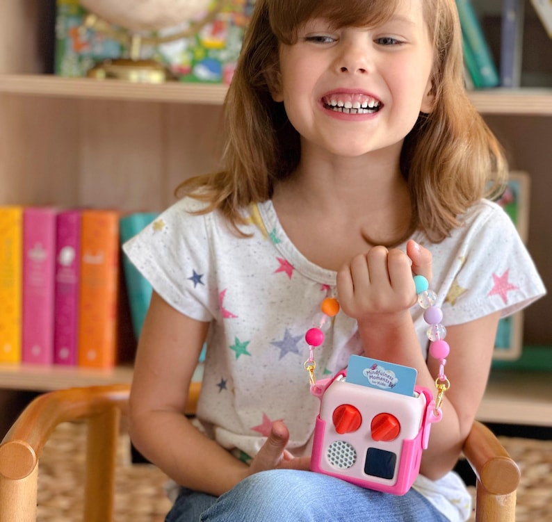 May include: A child holds a pink and white toy radio with red knobs and a small screen. The radio has a colourful beaded strap. The child wears a white t-shirt with colourful star graphics.