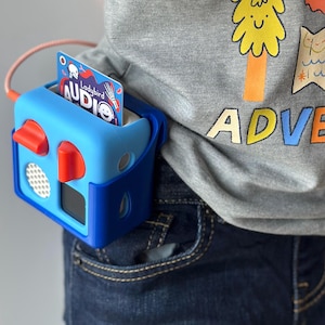 May include: A blue and red audio player with a card inserted, attached to a person's waist. The card reads "Ladybird Audio". The person is wearing a grey t-shirt with a tree graphic and the word "ADVENTURE".