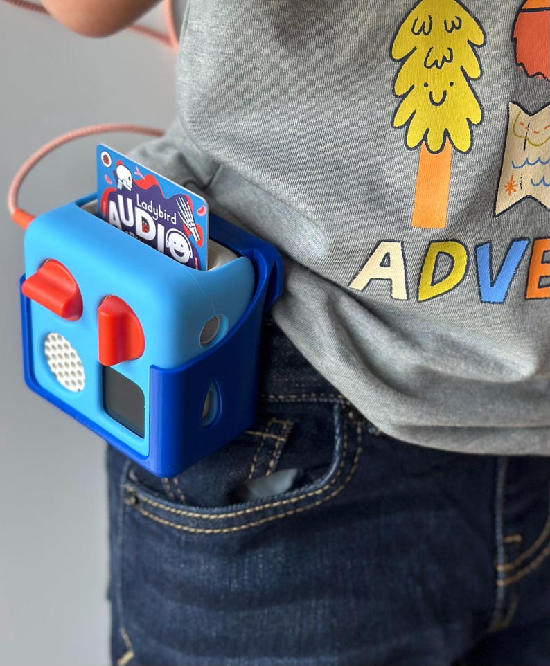 May include: A blue and red audio player with a card inserted, attached to a person's waist. The card reads "Ladybird Audio". The person is wearing a grey t-shirt with a tree graphic and the word "ADVENTURE".
