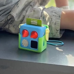 May include: A brightly coloured toy radio with a blue top, green sides, and red knobs. A card with the text "ANIMAL SONGS" is inserted in the top. The toy has a speaker and a black square. A blue cord is attached to the side.