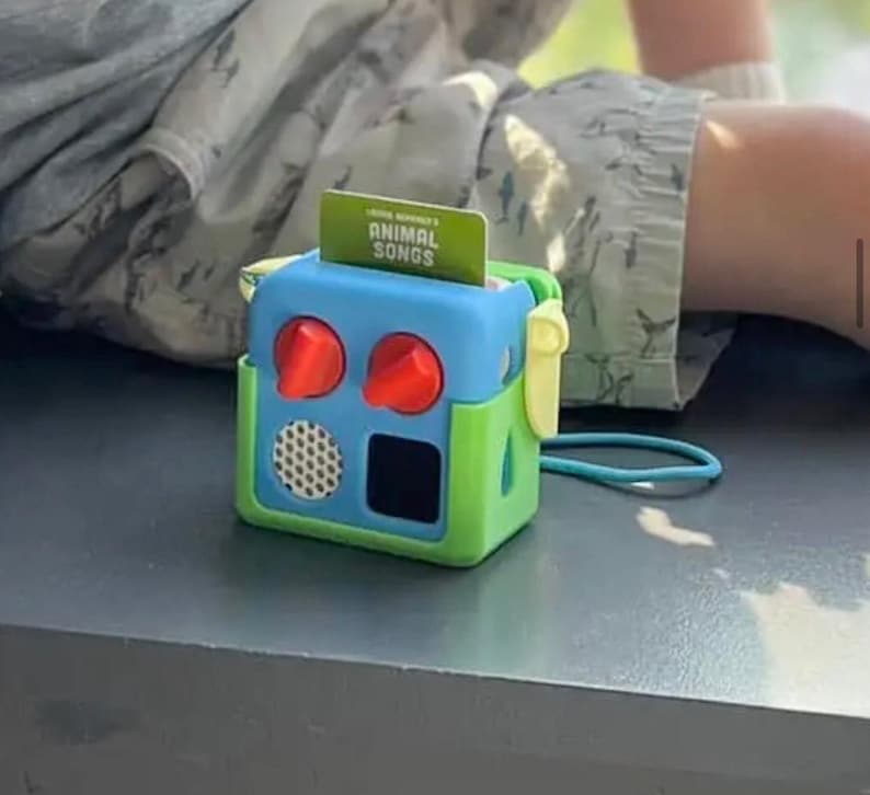 May include: A brightly coloured toy radio with a blue top, green sides, and red knobs. A card with the text "ANIMAL SONGS" is inserted in the top. The toy has a speaker and a black square. A blue cord is attached to the side.