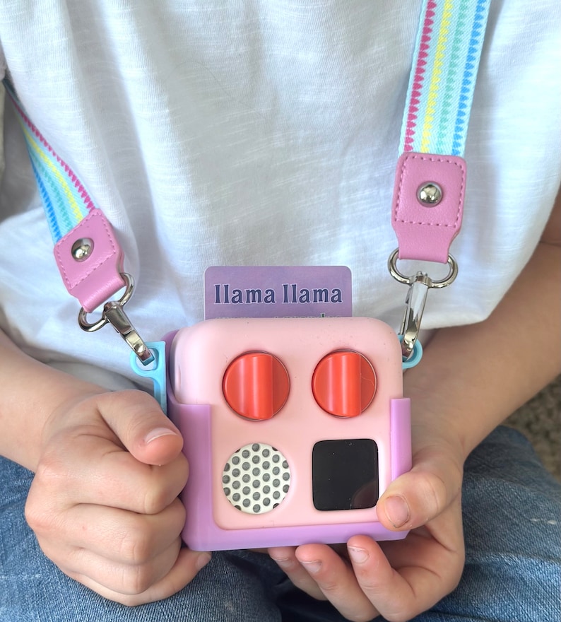 May include: A pink and purple toy with orange knobs, a black square, and a dotted circle. The toy has a strap with a colourful pattern and the text "Llama Llama" on a card.