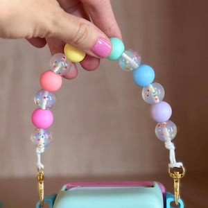 Peut inclure: Une sangle de perles color&eacute;es avec des perles aux couleurs pastel et des perles transparentes avec des confettis. La sangle est attach&eacute;e &agrave; un objet bleu clair avec des boutons rouges et des fermoirs dor&eacute;s.