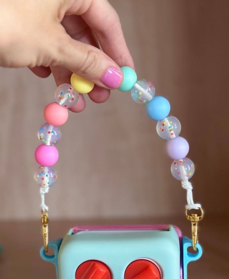 Peut inclure: Une sangle de perles color&eacute;es avec des perles aux couleurs pastel et des perles transparentes avec des confettis. La sangle est attach&eacute;e &agrave; un objet bleu clair avec des boutons rouges et des fermoirs dor&eacute;s.