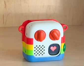 May include: A square-shaped electronic device case featuring a white front panel with two orange knobs, a speaker grille, and a small screen displaying a pink pixelated heart. The outer frame is coloured with horizontal stripes of red, yellow, green, and blue.
