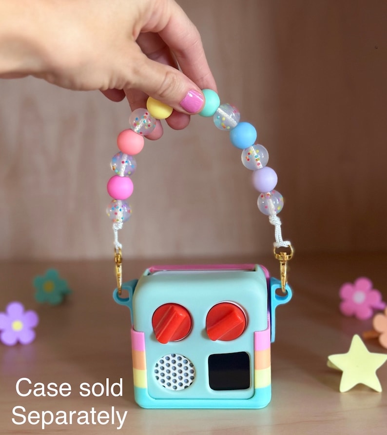 Peut inclure: Une bo&icirc;te &agrave; jouets bleu clair avec des boutons orange et un haut-parleur. Elle a un c&ocirc;t&eacute; ray&eacute; arc-en-ciel et une sangle perl&eacute;e avec des perles de couleur pastel. La sangle est tenue par une main. Le texte "Case sold Separately" est en bas.