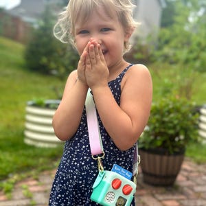 May include: A young child wearing a navy blue floral dress, holding a mint green and red toy camera purse with a rainbow strap. The purse has the text "Row Your Boat" on a small card. The child is smiling.