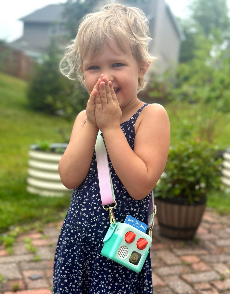 May include: A young child wearing a navy blue floral dress, holding a mint green and red toy camera purse with a rainbow strap. The purse has the text "Row Your Boat" on a small card. The child is smiling.