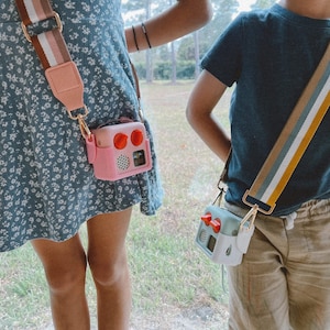 May include: Two children wearing crossbody bags with colourful straps. One bag is pink with red accents, the other is mint green with red accents. Both bags have a retro radio design and are attached to the straps with gold-coloured hardware.