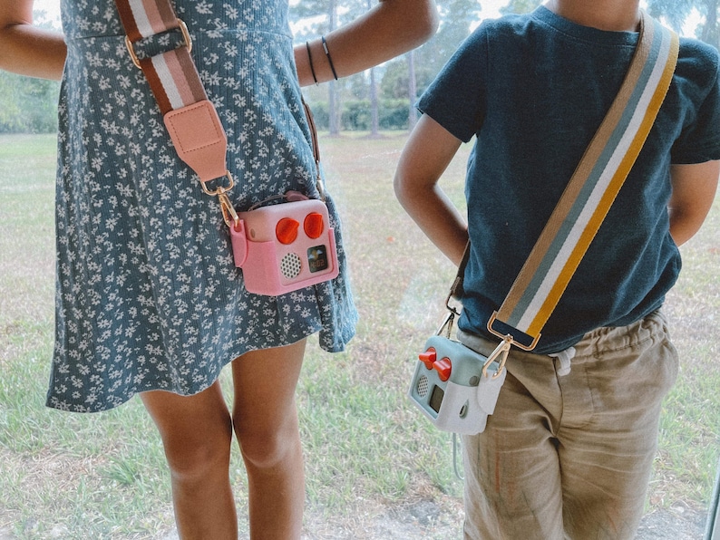 May include: Two children wearing crossbody bags with colourful straps. One bag is pink with red accents, the other is mint green with red accents. Both bags have a retro radio design and are attached to the straps with gold-coloured hardware.