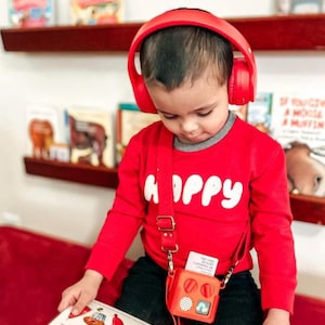 May include: A child wearing red headphones and a red long-sleeved top with the word "HAPPY" in white. The child is reading a book with colourful illustrations and has a small red square bag with a strap.