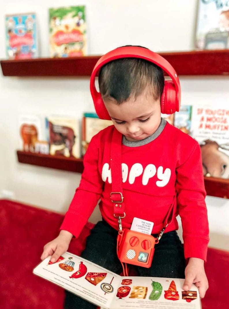 May include: A child wearing red headphones and a red long-sleeved top with the word "HAPPY" in white. The child is reading a book with colourful illustrations and has a small red square bag with a strap.