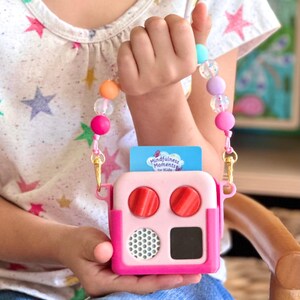 May include: A pink and white toy featuring two red circular buttons, a small black square, and a speaker grill. It has a beaded strap with colourful beads and a card that reads "Mindfulness Moments for Kids."
