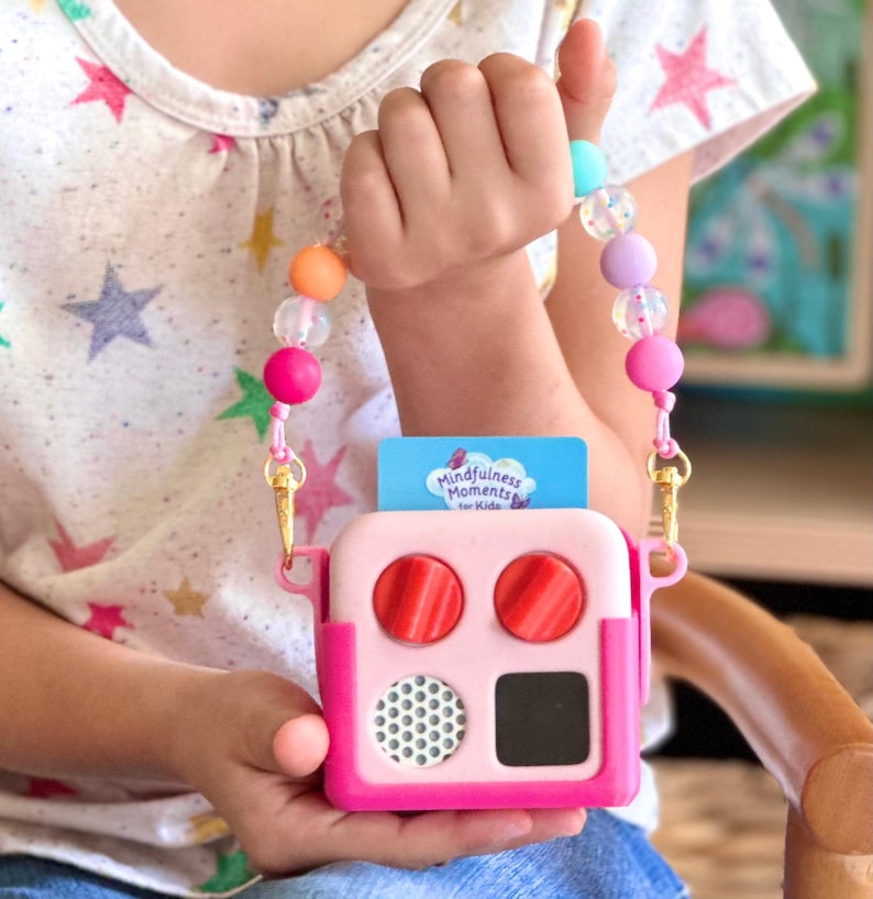May include: A pink and white toy featuring two red circular buttons, a small black square, and a speaker grill. It has a beaded strap with colourful beads and a card that reads "Mindfulness Moments for Kids."