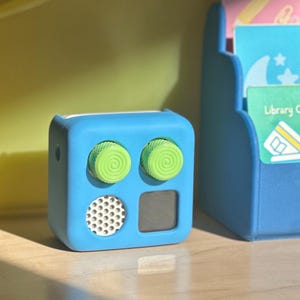 May include: A small, blue, cube-shaped device with two green, spiral-patterned knobs. It has a speaker and a small, dark square. A blue shelf with cards is in the background.