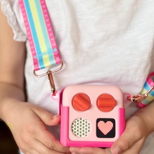 May include: A pink and magenta square crossbody bag with a colourful striped strap. The bag has two orange dials, a speaker, and a pixelated heart design. The strap has pink hearts and rainbow stripes.