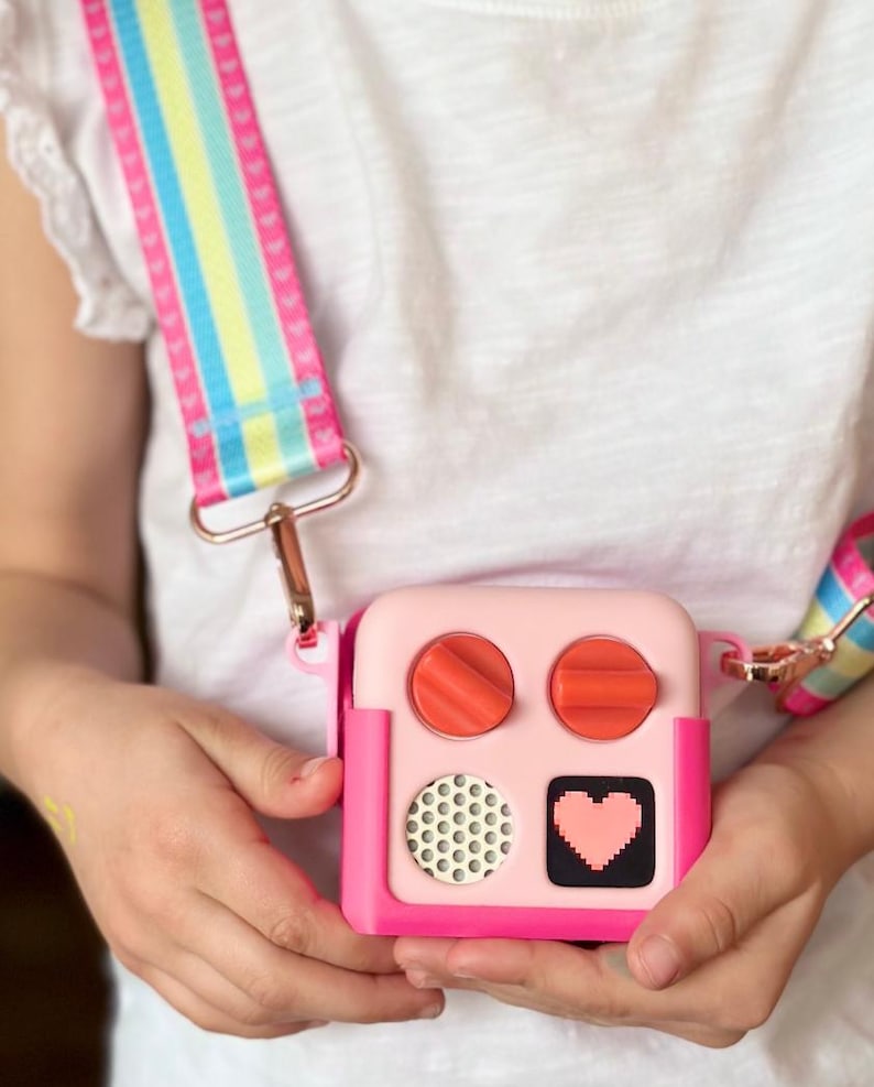 May include: A pink and magenta square crossbody bag with a colourful striped strap. The bag has two orange dials, a speaker, and a pixelated heart design. The strap has pink hearts and rainbow stripes.