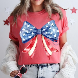 May include: A coral-colored t-shirt featuring a large bow design. The bow has a blue center with white stars and red and white striped ribbons, evoking an American flag theme. The shirt is a casual style.