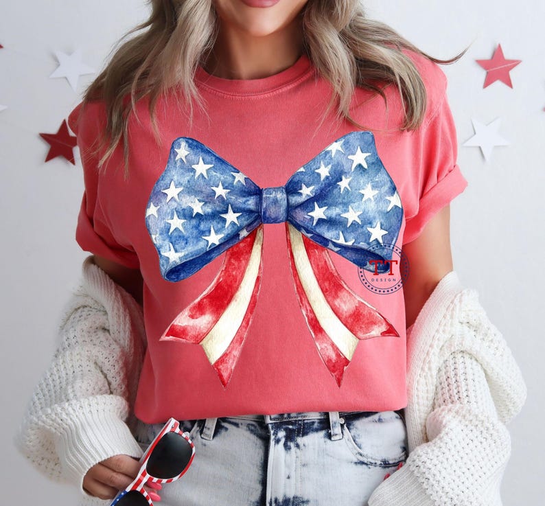 May include: A coral-colored t-shirt featuring a large bow design. The bow has a blue center with white stars and red and white striped ribbons, evoking an American flag theme. The shirt is a casual style.