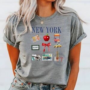 Op de afbeelding: Grijs T-shirt met de tekst "NEW YORK" in blauw. Het shirt heeft verschillende iconen met het thema New York City, waaronder een rode appel, een gele taxi, een hotdogkraam en een Broadway-bord.