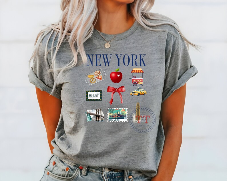 Op de afbeelding: Grijs T-shirt met de tekst "NEW YORK" in blauw. Het shirt heeft verschillende iconen met het thema New York City, waaronder een rode appel, een gele taxi, een hotdogkraam en een Broadway-bord.