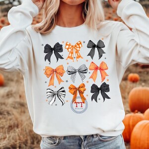 Pu&ograve; includere: Felpa bianca con un design grafico di fiocchi arancioni, neri e bianchi. I fiocchi sono disposti in una griglia e hanno uno stile vintage disegnato a mano. Il testo "TT Design" &egrave; stampato nella parte inferiore centrale della felpa.