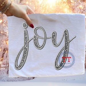 Könnte beinhalten: Weißer Stoff mit dem Wort "Joy" in Schreibschrift gestickt. Die Buchstaben sind mit einem silbernen Glitzerdesign umrandet. Ein rot-blaues Logo ist unten rechts sichtbar. Der Hintergrund ist eine Winterlandschaft mit schneebedeckten Bäumen.