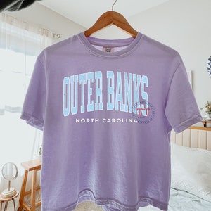May include: A lavender t-shirt with a blue graphic that says "Outer Banks North Carolina" with a small red circle logo.