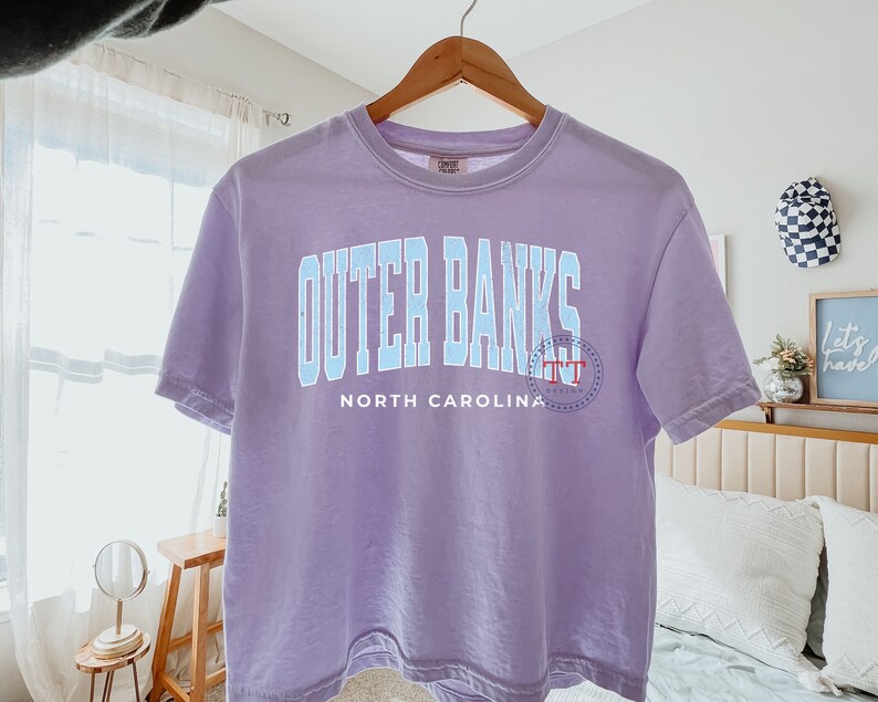 May include: A lavender t-shirt with a blue graphic that says "Outer Banks North Carolina" with a small red circle logo.