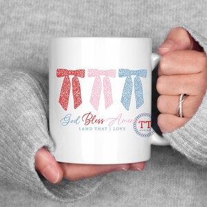 Puede incluir: Taza de cer&aacute;mica blanca con un dise&ntilde;o de lazos rojo, rosa y azul. El texto "God Bless America Land That I Love" est&aacute; impreso en la taza.