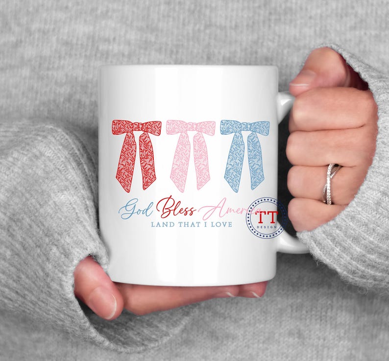 Puede incluir: Taza de cer&aacute;mica blanca con un dise&ntilde;o de lazos rojo, rosa y azul. El texto "God Bless America Land That I Love" est&aacute; impreso en la taza.