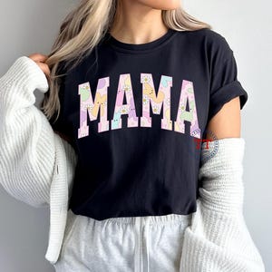 Puede incluir: Camiseta negra con la palabra "MAMA" en letras rosas. Las letras est&aacute;n rellenas con un patr&oacute;n pastel de conejos y flores.