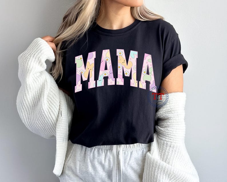 Puede incluir: Camiseta negra con la palabra "MAMA" en letras rosas. Las letras est&aacute;n rellenas con un patr&oacute;n pastel de conejos y flores.